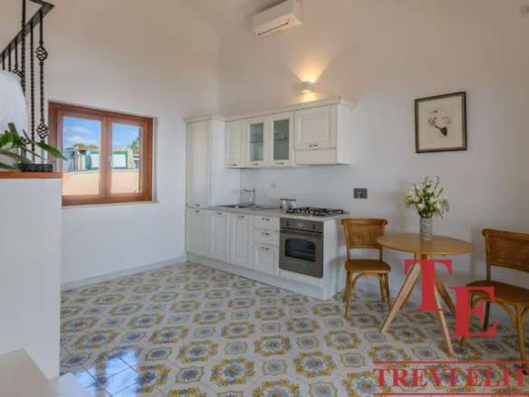 Уютная кухня с белой мебелью и деревянными окнами, светлый интерьер, стильный стол и два стула, свежие цветы в вазе создают приятную атмосферу для жизни и отдыха. Название агентства "Треви Элит" на изображении.