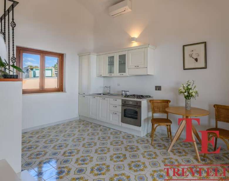 Уютная кухня с белой мебелью и деревянными окнами, светлый интерьер, стильный стол и два стула, свежие цветы в вазе создают приятную атмосферу для жизни и отдыха. Название агентства "Треви Элит" на изображении.