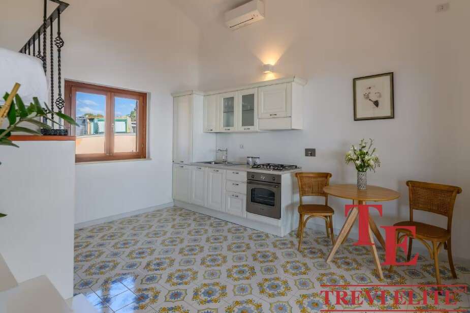 Уютная кухня с белой мебелью и деревянными окнами, светлый интерьер, стильный стол и два стула, свежие цветы в вазе создают приятную атмосферу для жизни и отдыха. Название агентства "Треви Элит" на изображении.