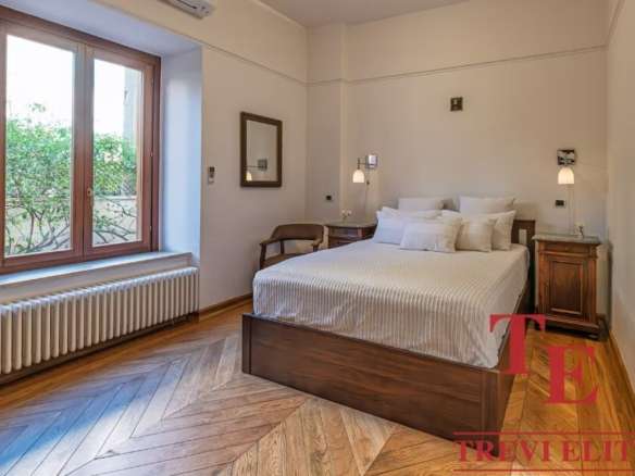 Элегантная спальня в стильной классической обстановке с большим окном и уютной атмосферой. В интерьере используются теплые древесные тона и современные светильники для комфорта и эстетики.