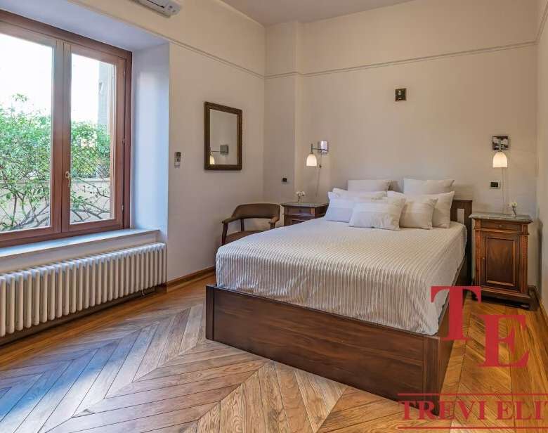 Элегантная спальня в стильной классической обстановке с большим окном и уютной атмосферой. В интерьере используются теплые древесные тона и современные светильники для комфорта и эстетики.