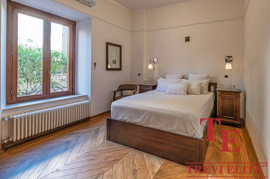 Элегантная спальня в стильной классической обстановке с большим окном и уютной атмосферой. В интерьере используются теплые древесные тона и современные светильники для комфорта и эстетики.