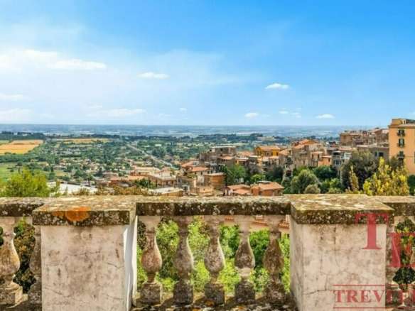 Вид на живописный итальянский пейзаж с балкона, охватывающий городские улицы, зелёные насаждения и сельскую местность на фоне голубого неба.