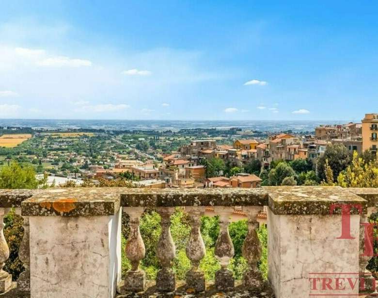 Вид на живописный итальянский пейзаж с балкона, охватывающий городские улицы, зелёные насаждения и сельскую местность на фоне голубого неба.