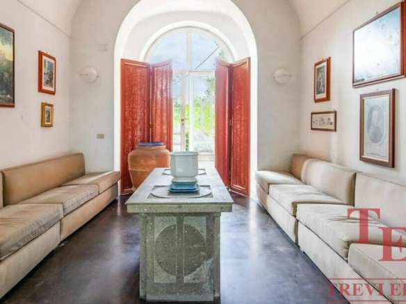 Элегантная гостиная в роскошной итальянской недвижимости с светлыми диванами, искусственными картинами и ярким светом, входом через арку с деревянными дверями.