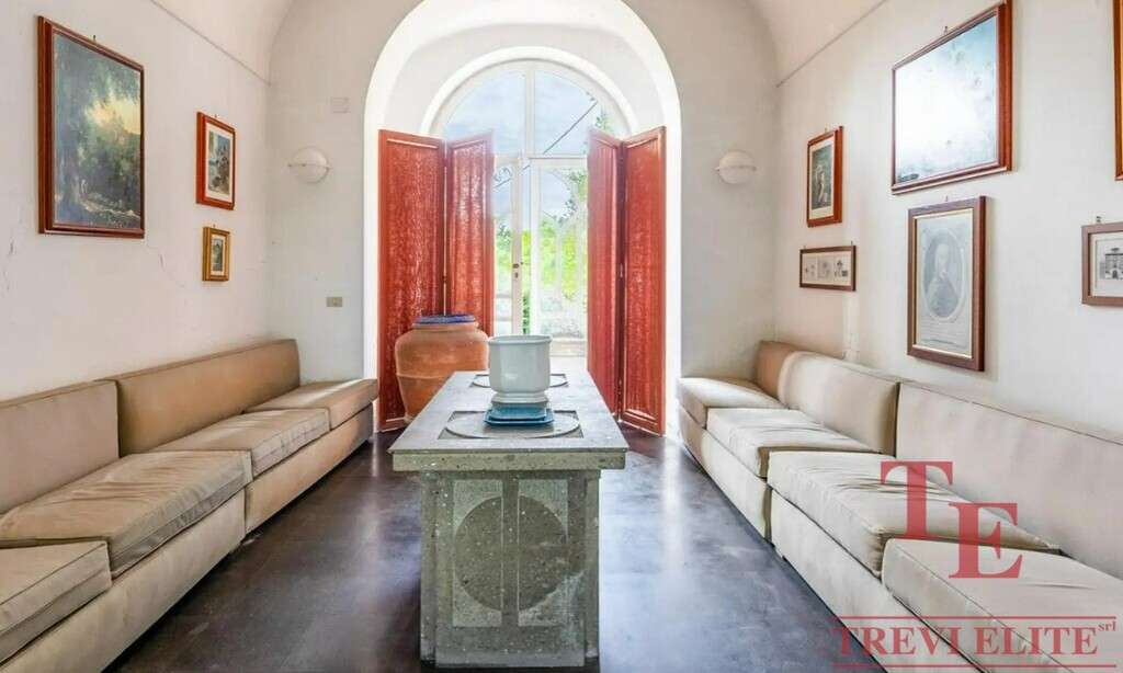 Элегантная гостиная в роскошной итальянской недвижимости с светлыми диванами, искусственными картинами и ярким светом, входом через арку с деревянными дверями.