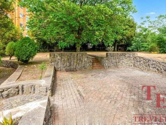 Уютная зона отдыха с зелеными деревьями и мощеной дорожкой, окруженная декоративными кустарниками и каменными стенами, идеально подходит для комфортных прогулок и отдыха на свежем воздухе возле элитной недвижимости.