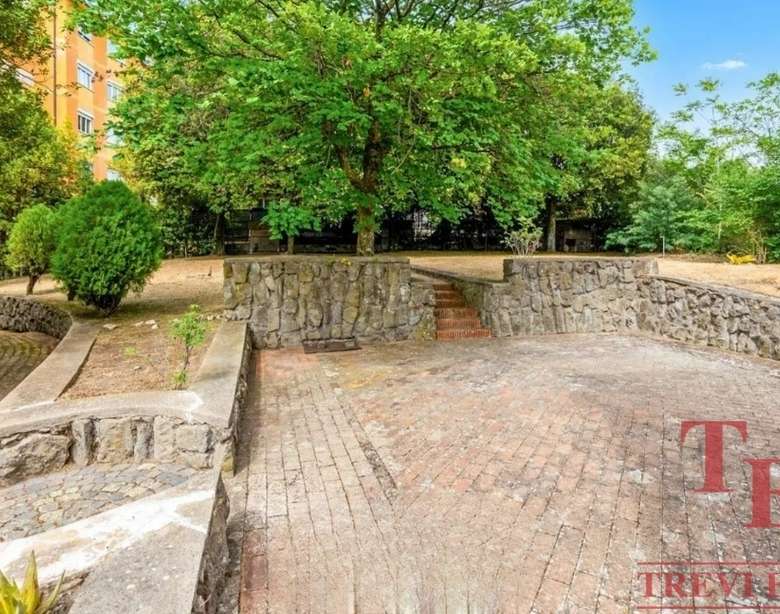 Уютная зона отдыха с зелеными деревьями и мощеной дорожкой, окруженная декоративными кустарниками и каменными стенами, идеально подходит для комфортных прогулок и отдыха на свежем воздухе возле элитной недвижимости.