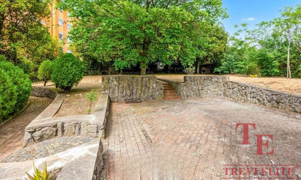 Уютная зона отдыха с зелеными деревьями и мощеной дорожкой, окруженная декоративными кустарниками и каменными стенами, идеально подходит для комфортных прогулок и отдыха на свежем воздухе возле элитной недвижимости.