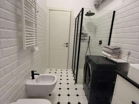 Современная благоустроенная ванная комната с белой кафельной отделкой, душевой кабиной, стиральной машиной и мебелью, создающая стильное и уютное пространство для комфорта.