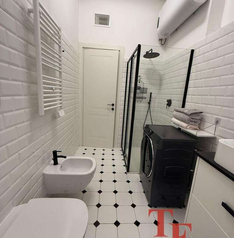 Современная благоустроенная ванная комната с белой кафельной отделкой, душевой кабиной, стиральной машиной и мебелью, создающая стильное и уютное пространство для комфорта.