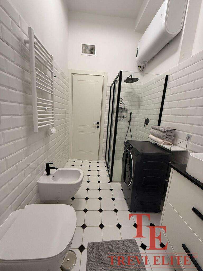 Современная благоустроенная ванная комната с белой кафельной отделкой, душевой кабиной, стиральной машиной и мебелью, создающая стильное и уютное пространство для комфорта.