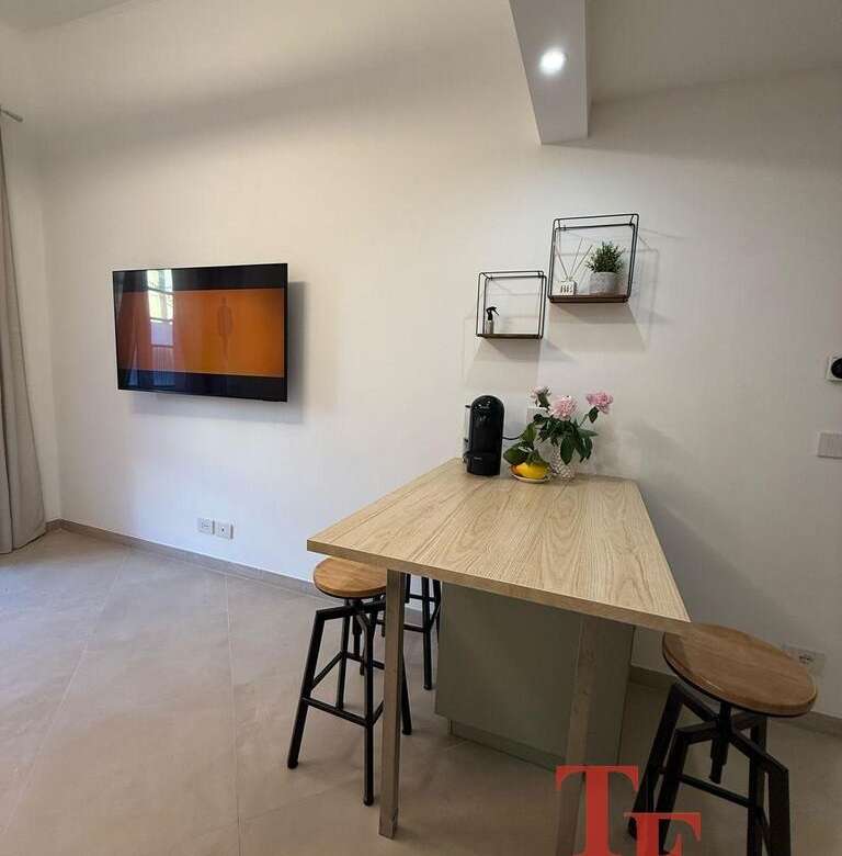 Современная уютная кухня с деревянным столом и барными стульями, телевизором и декоративными полками, создающими стильное пространство для отдыха и общения.