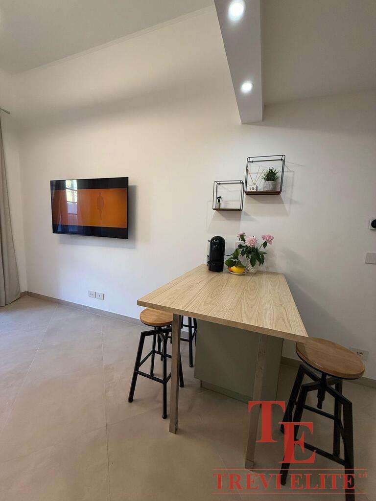 Современная уютная кухня с деревянным столом и барными стульями, телевизором и декоративными полками, создающими стильное пространство для отдыха и общения.