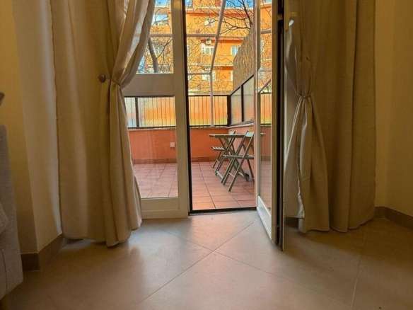 Изображение балкона с открытой дверью, вид на улицу с жилым домом и деревьями, внутри светлая комната с занавесками.