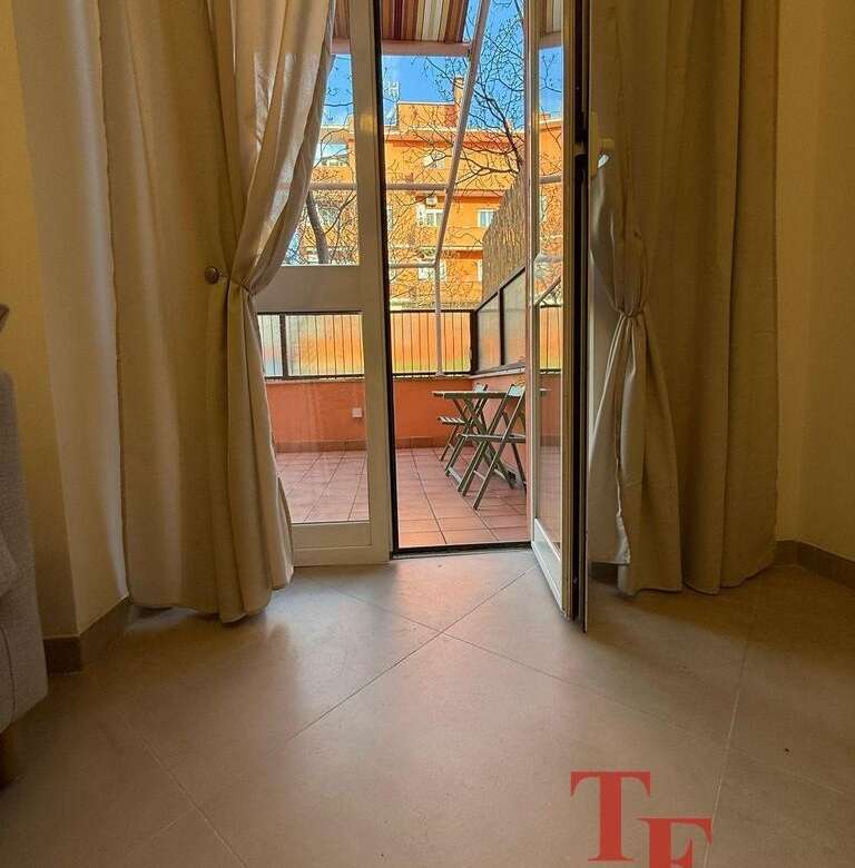 Изображение балкона с открытой дверью, вид на улицу с жилым домом и деревьями, внутри светлая комната с занавесками.