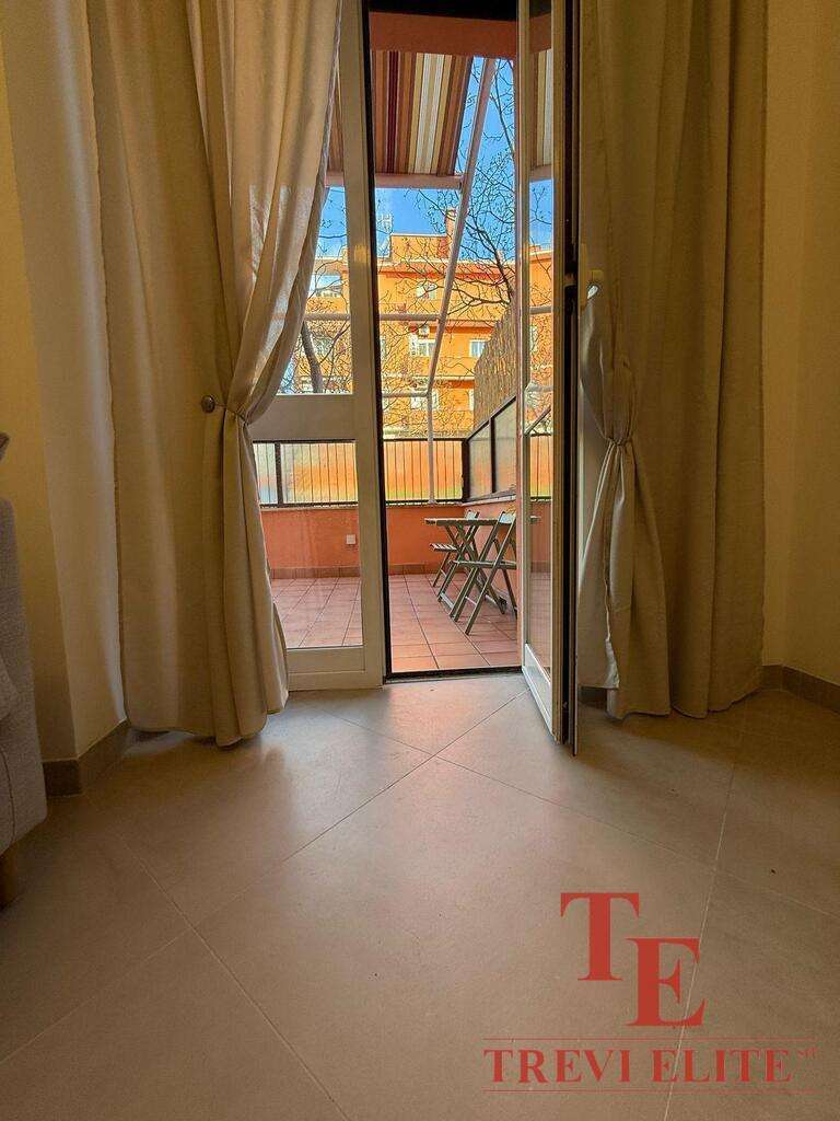 Изображение балкона с открытой дверью, вид на улицу с жилым домом и деревьями, внутри светлая комната с занавесками.