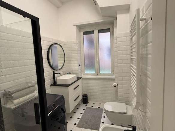 Современная дизайнерская ванная комната с белой плиткой, стиральной машиной, раковиной и теплым полотенцесушителем, светлое окно, оформленная в стиле лаконичного минимализма.