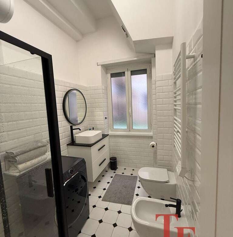 Современная дизайнерская ванная комната с белой плиткой, стиральной машиной, раковиной и теплым полотенцесушителем, светлое окно, оформленная в стиле лаконичного минимализма.