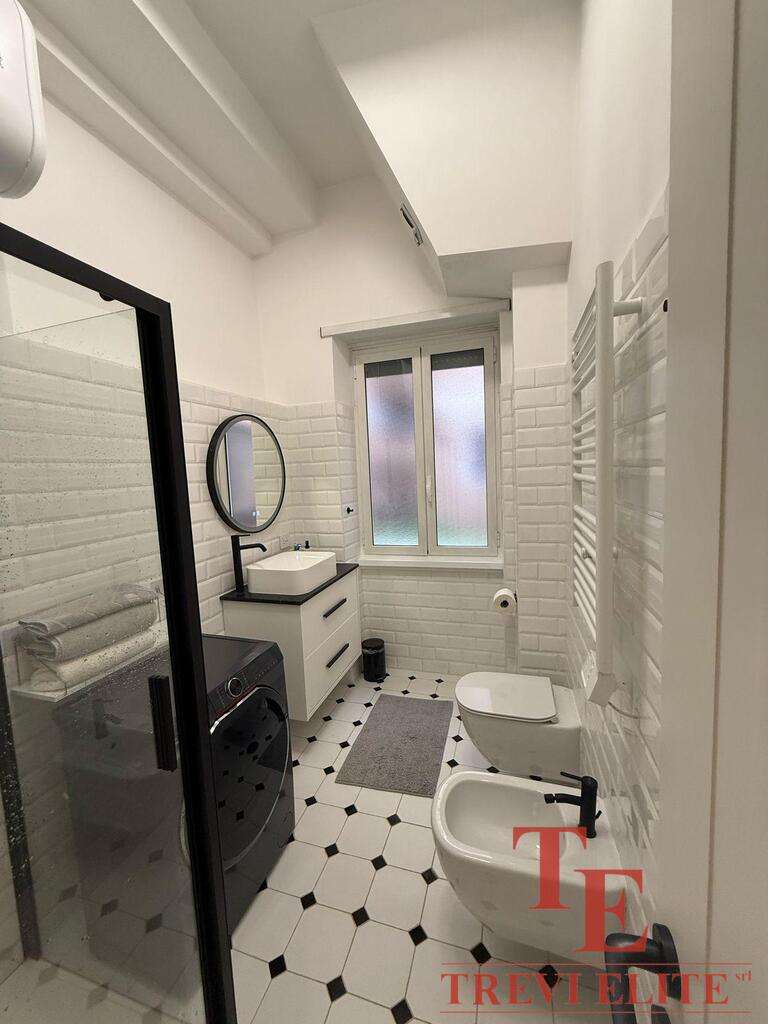 Современная дизайнерская ванная комната с белой плиткой, стиральной машиной, раковиной и теплым полотенцесушителем, светлое окно, оформленная в стиле лаконичного минимализма.