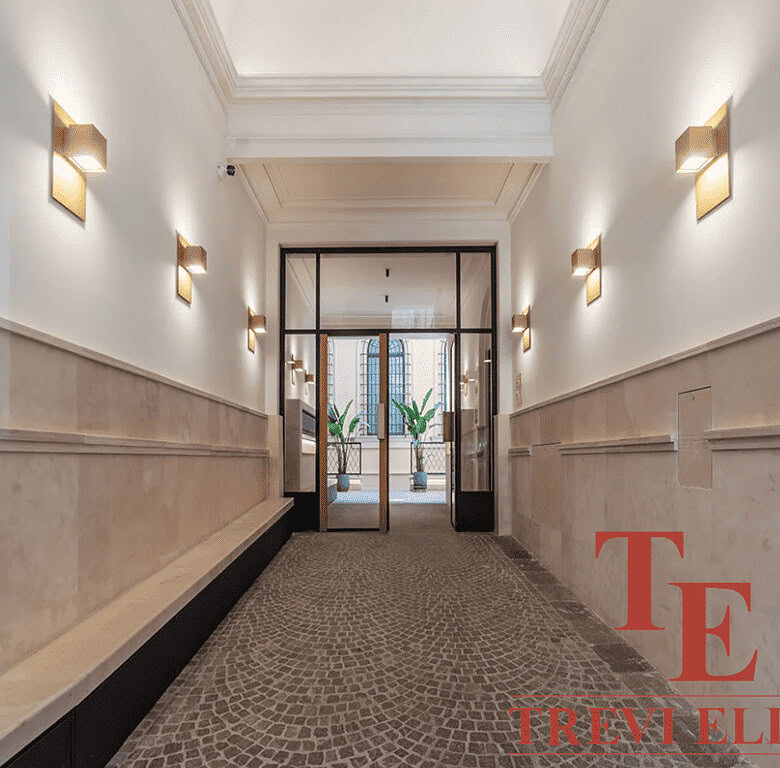 Элегантный коридор в офисном здании с современным дизайном, белыми стенами, множеством светильников и стеклянной дверью, ведущей на прогулочный балкон, создавая атмосферу роскоши и стиля.