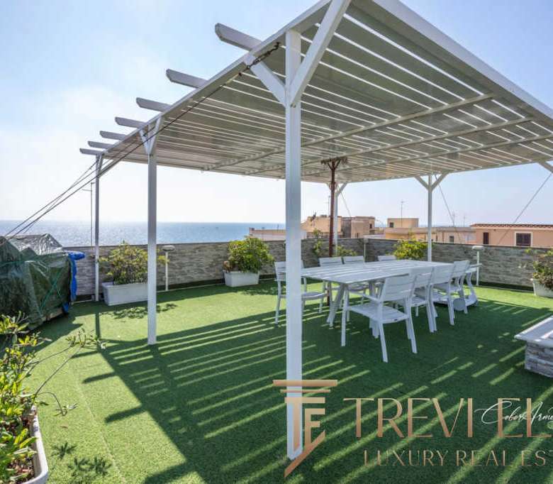 Уютная терраса с солнечной защитой, цветущими растениями и видом на море, идеально подходит для отдыха и приёма гостей на роскошной недвижимости в Италии.