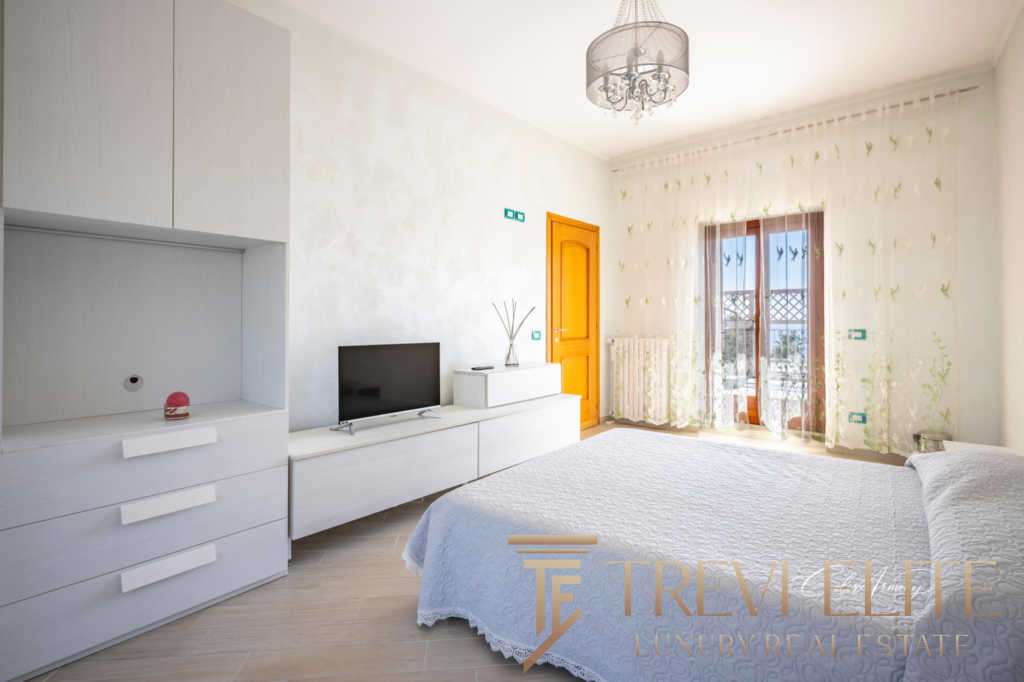 Роскошная спальня с чистым дизайном и уютной атмосферой, большие окна с шторками и французским балконом, светлый интерьер, современные элементы, подходящая для элитной недвижимости.