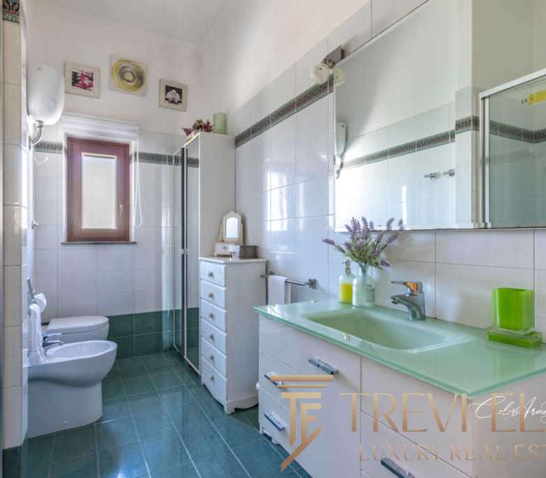 Современная ванная комната с белыми стенами и зеленым акцентным умывальником, большое зеркало и аккуратная мебель, создающие атмосферу уюта и роскоши в престижной недвижимости.