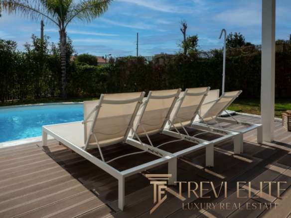 Высококлассные шезлонги у бассейна на террасе роскошного загородного дома с видом на сад и пальмы. Идеальное место для отдыха и релаксации в эксклюзивной недвижимости.