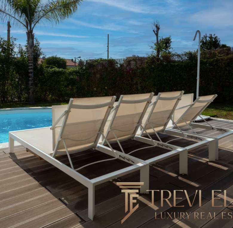 Высококлассные шезлонги у бассейна на террасе роскошного загородного дома с видом на сад и пальмы. Идеальное место для отдыха и релаксации в эксклюзивной недвижимости.