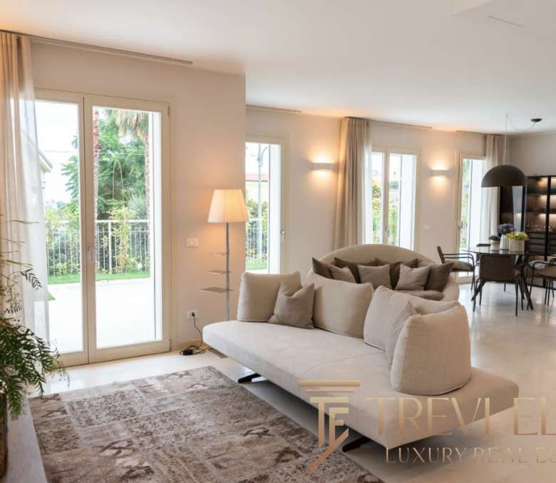 Элитная гостиная с панорамными окнами, современным дизайном, уютной обстановкой и видом на зеленый сад, идеально для отдыха и встреч.