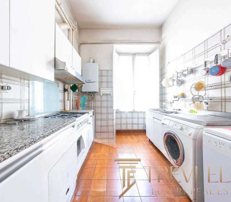 Богатая кухня в уютной квартире с белыми шкафами и стиральной машиной, яркие чашки и посуда в современном стиле, просторное освещенное помещение с окнами и теплым полом.