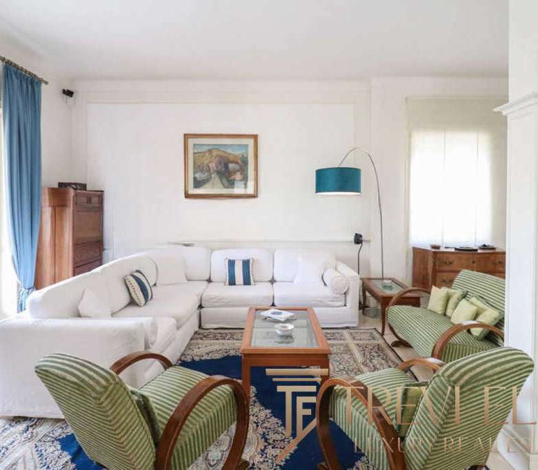 Просторная гостиная в роскошном доме с белой мебелью, зелеными креслами и уютными элементами интерьера, оформленными в светлых тонах, создает атмосферу уюта и элегантности.