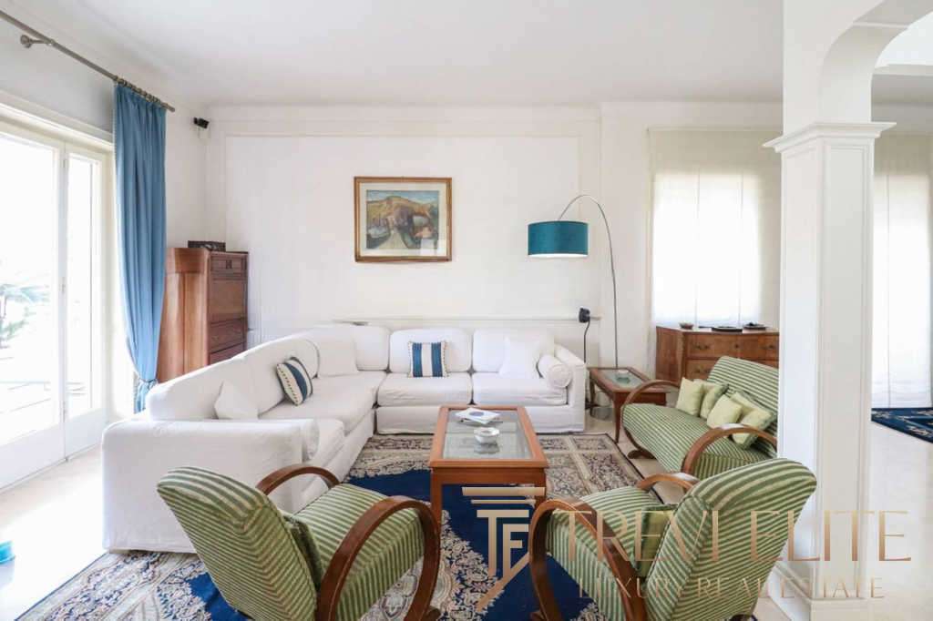 Просторная гостиная в роскошном доме с белой мебелью, зелеными креслами и уютными элементами интерьера, оформленными в светлых тонах, создает атмосферу уюта и элегантности.