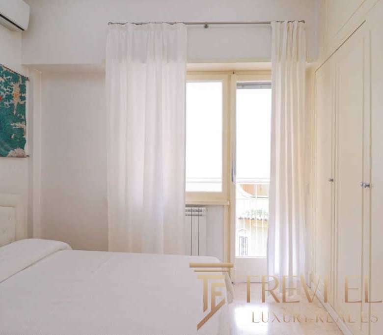 Роскошная спальня с белыми стенами и большими окнами, украшенными светлыми шторами, светлое мебельное оформление, создающее комфортную атмосферу для высококлассной недвижимости.