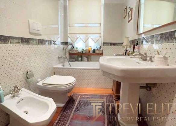 Bадовая комната с унитазом, биде и раковиной в домашнем интерьере, светлые стены с декоративной плиткой, окно с занавесками и зеркалом, включает элементы роскоши и уюта, идеально для реализации эксклюзивной недвижимости.