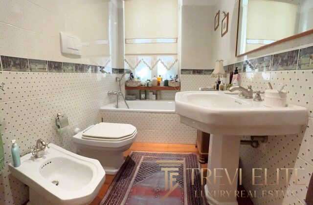 Bадовая комната с унитазом, биде и раковиной в домашнем интерьере, светлые стены с декоративной плиткой, окно с занавесками и зеркалом, включает элементы роскоши и уюта, идеально для реализации эксклюзивной недвижимости.