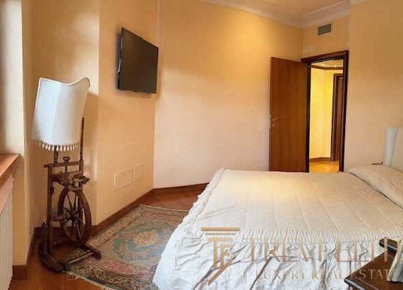 Уютная спальня с мягкой кроватью, телевизором и классическим украшением в стиле антика, светлые стены, панорамное окно, создающее теплую атмосферу для комфортного отдыха.