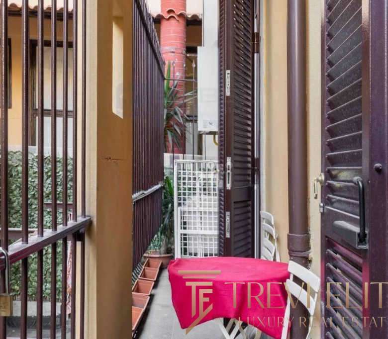 Узкий балкон с металлическими решетками и шторками, уютное место для отдыха с мягким креслом и ярким покрывалом, окруженный жилыми домами и зеленью, идеально для ценителей роскошных итальянских и международных элитных недвижимых объектов.