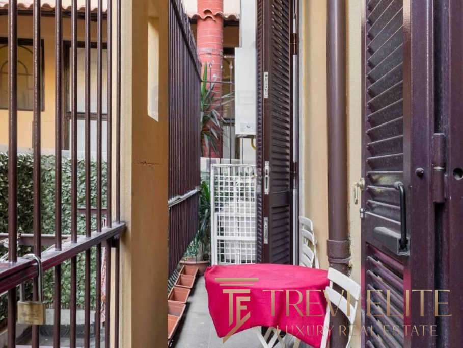 Узкий балкон с металлическими решетками и шторками, уютное место для отдыха с мягким креслом и ярким покрывалом, окруженный жилыми домами и зеленью, идеально для ценителей роскошных итальянских и международных элитных недвижимых объектов.