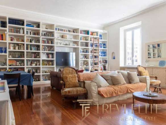 Современная гостиная с множеством книг в белой библиотеке, уютной мебелью и большим окном, пропускающим естественный свет. Идеальное место для отдыха и чтения в престижной недвижимости.