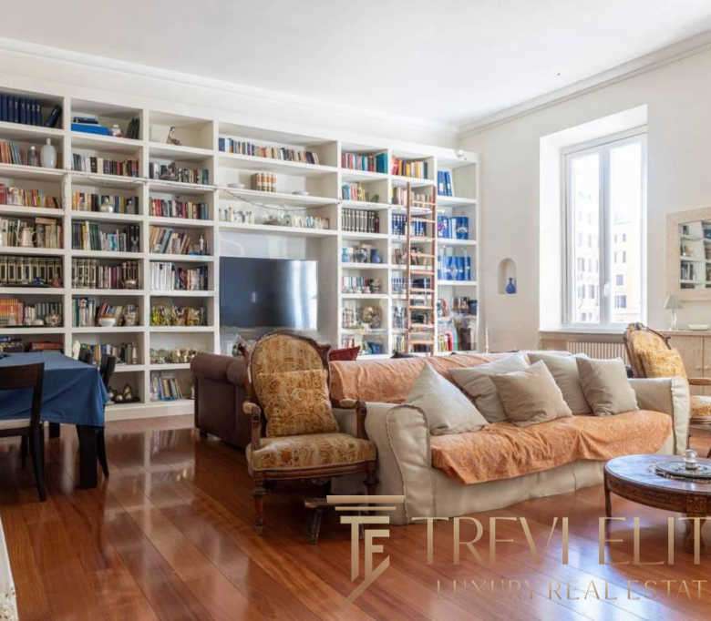 Современная гостиная с множеством книг в белой библиотеке, уютной мебелью и большим окном, пропускающим естественный свет. Идеальное место для отдыха и чтения в престижной недвижимости.