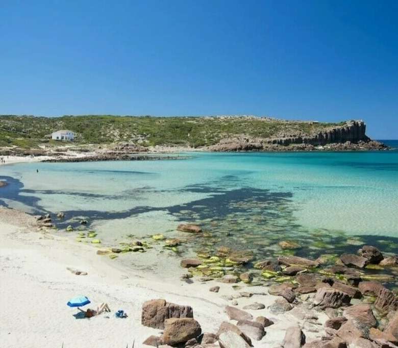 Великолепный пляж на берегу с прозрачной бирюзовой водой, песчаным побережьем и скалами, окруженными зеленью и бескрайним синим небом, идеальное место для отдыха и наслаждения природой.