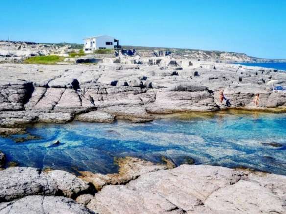 Живописный берег с прозрачной голубой водой, скалами и современным домом на фоне, идеально подходит для элитной недвижимости и отдыха на побережье Средиземноморья.