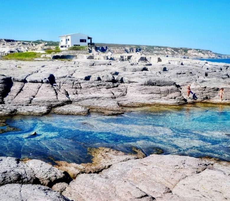 Живописный берег с прозрачной голубой водой, скалами и современным домом на фоне, идеально подходит для элитной недвижимости и отдыха на побережье Средиземноморья.