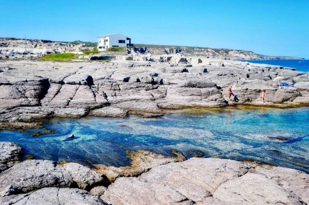 Живописный берег с прозрачной голубой водой, скалами и современным домом на фоне, идеально подходит для элитной недвижимости и отдыха на побережье Средиземноморья.