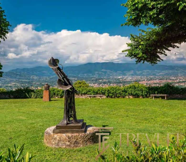 Современная скульптура на лужайке с панорамным видом на горы и город, зеленые деревья и чистое небо, расположен в элитной недвижимостной зоне Италии, эксклюзивное агентство Треви Элит.