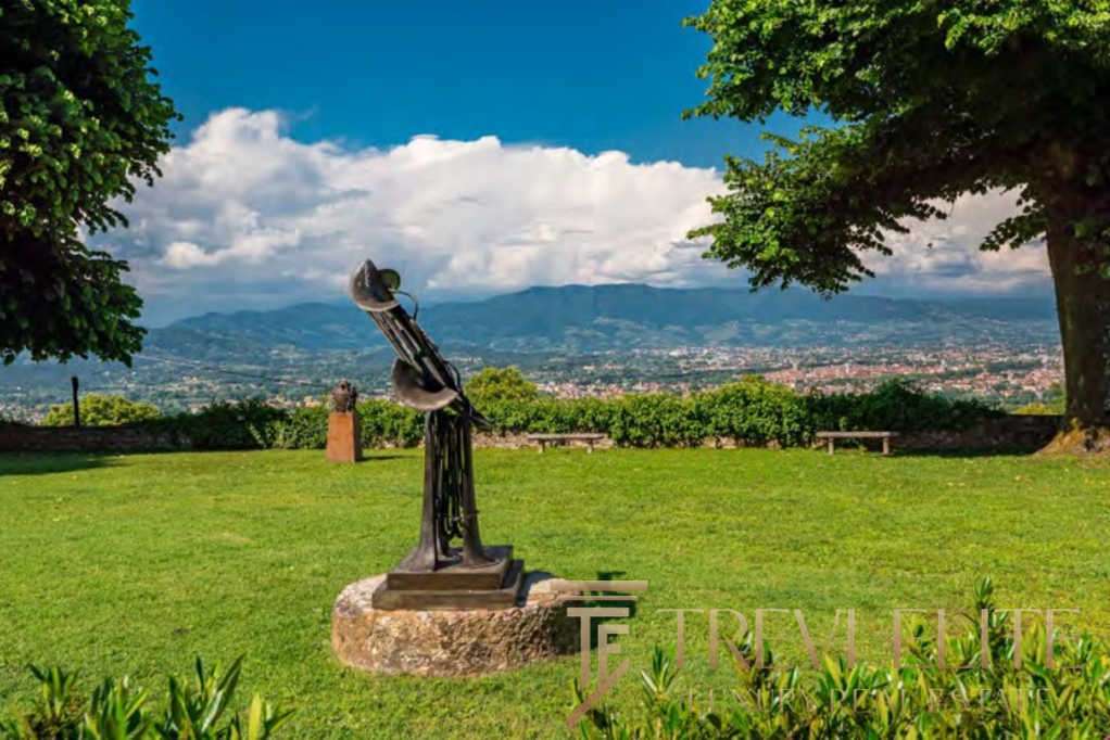 Современная скульптура на лужайке с панорамным видом на горы и город, зеленые деревья и чистое небо, расположен в элитной недвижимостной зоне Италии, эксклюзивное агентство Треви Элит.