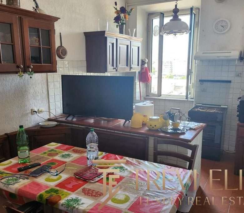 Обеденный стол в жилой кухне с яркой скатертью и напитками, рядом телевизор, открытое окно, создающее уютную атмосферу домашнего уюта и высокого уровня жизни.