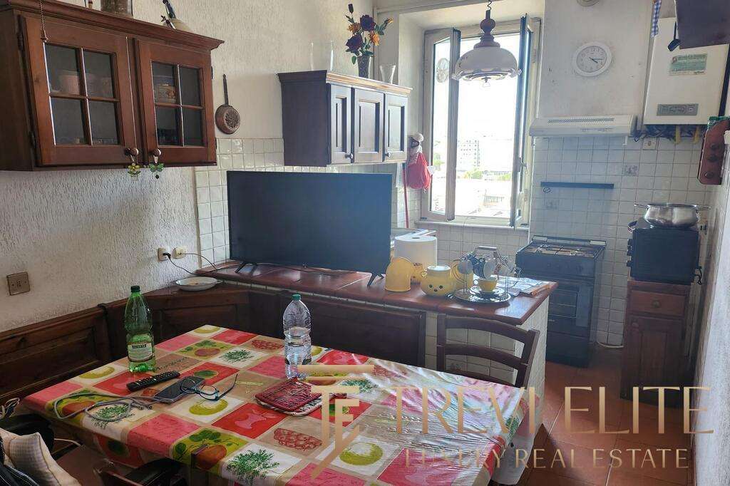 Обеденный стол в жилой кухне с яркой скатертью и напитками, рядом телевизор, открытое окно, создающее уютную атмосферу домашнего уюта и высокого уровня жизни.